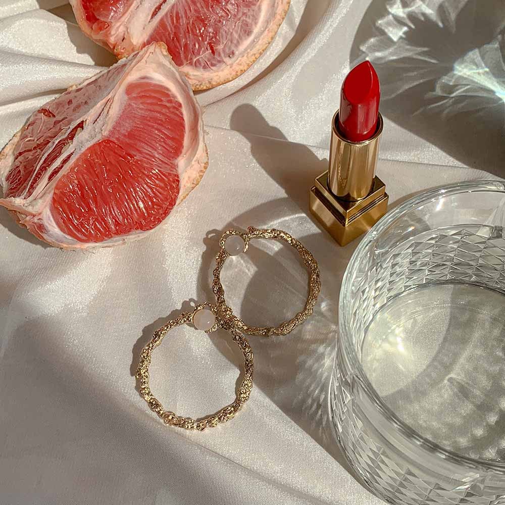Gold hoops beside a grapefruit, a lipstick and a glass