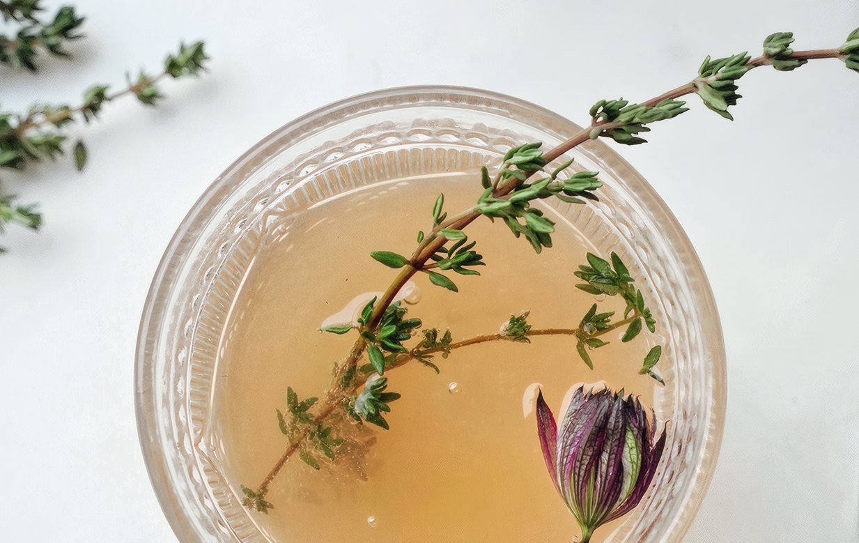 A glass bowl of tea with a sprig of thyme and a flower bud