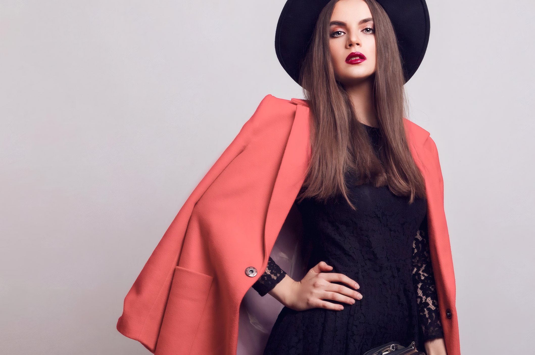 A model in a coral coat and a black hat on a pale grey sweep