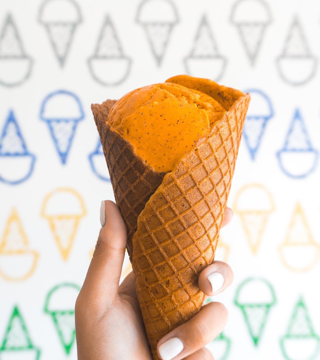 A waffle cone of orange sorbet held against a cone-print wall