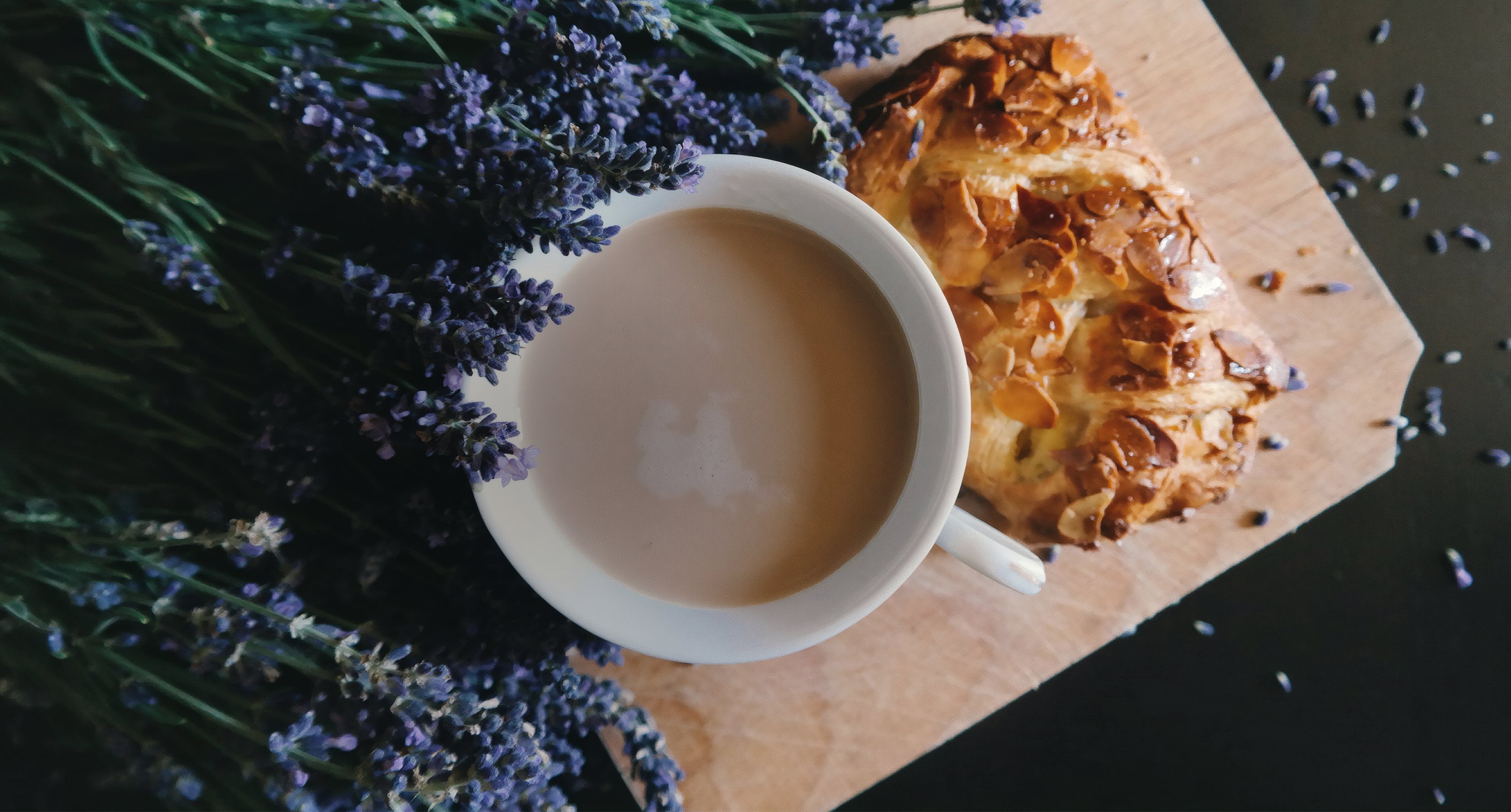 A latte and an almond pastry on a board beside lavender