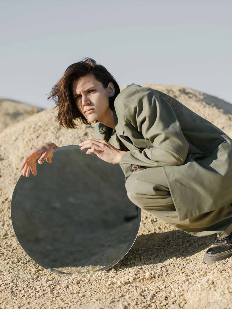 A woman crouching with a round mirror on the sand, from The Mirror