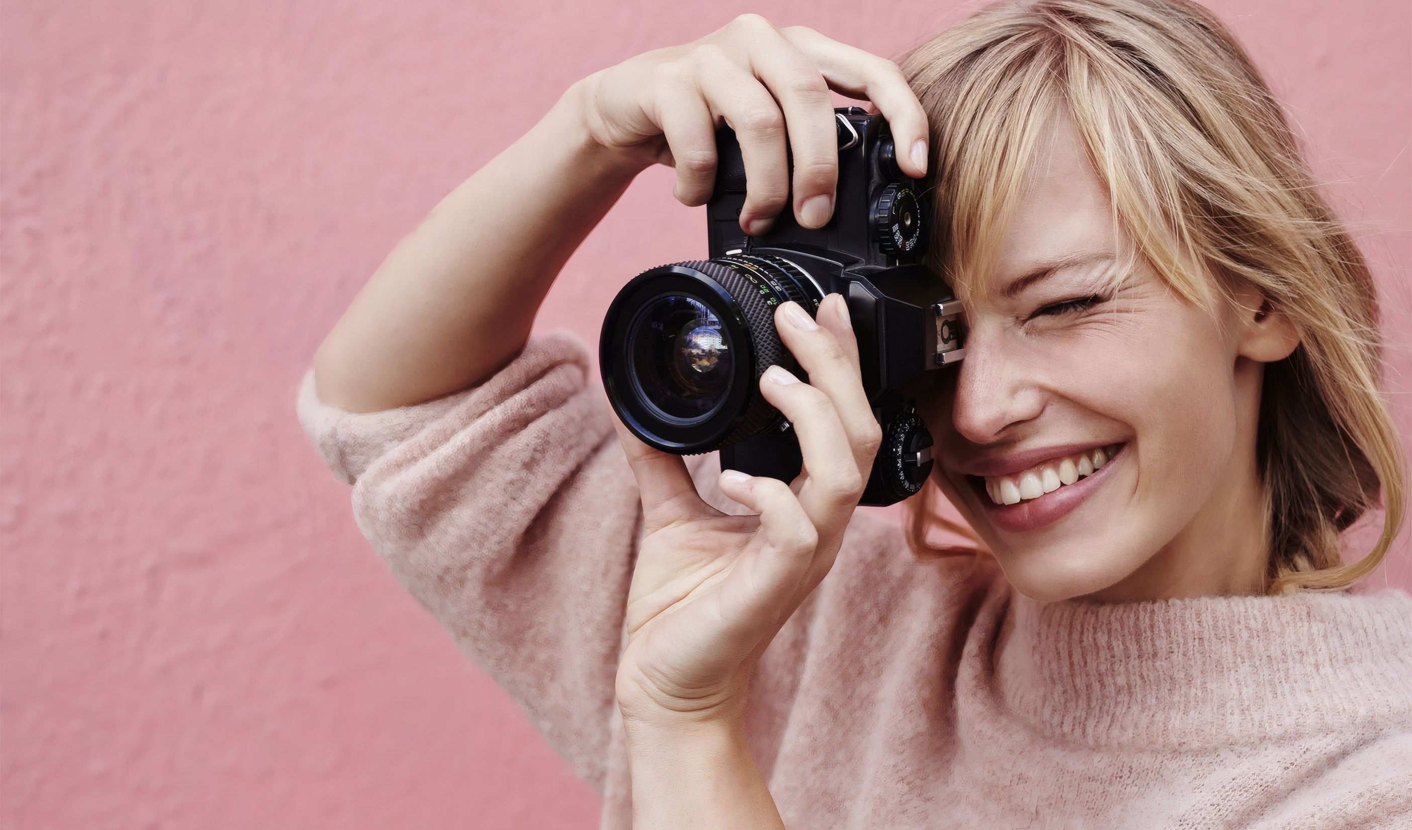 A smiling photography student with a camera against a pink wall
