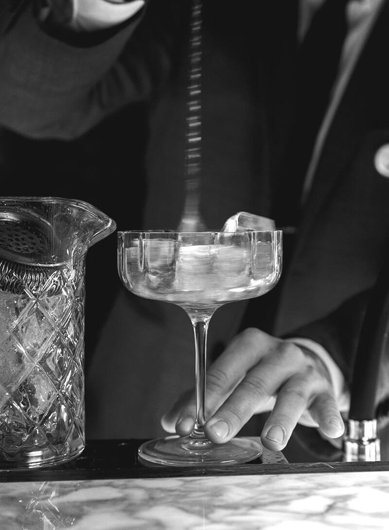 A bartender straining a drink into a coupe, in black and white