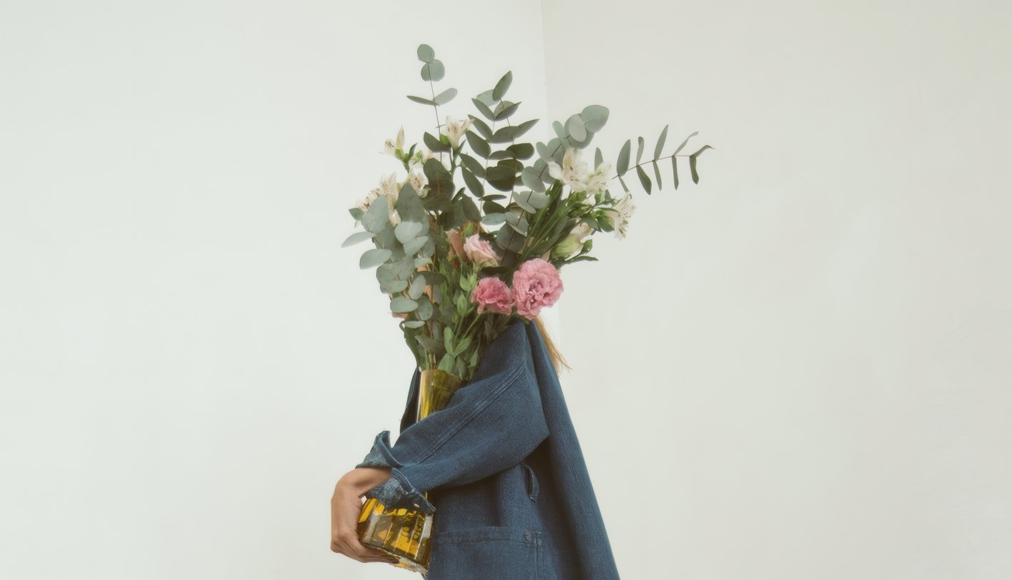 A person in an indigo chore coat holding a bouquet against a white wall