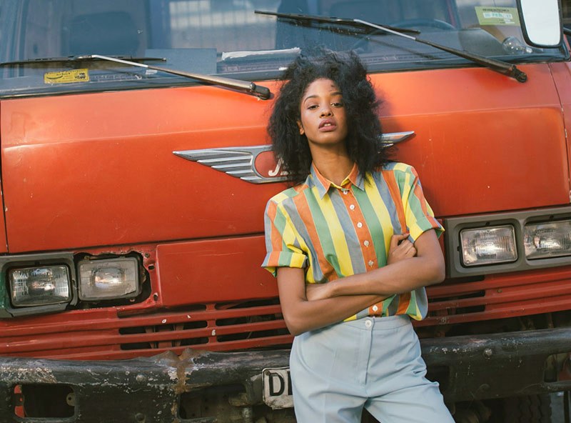 A woman in a striped camp shirt leaning on an orange truck