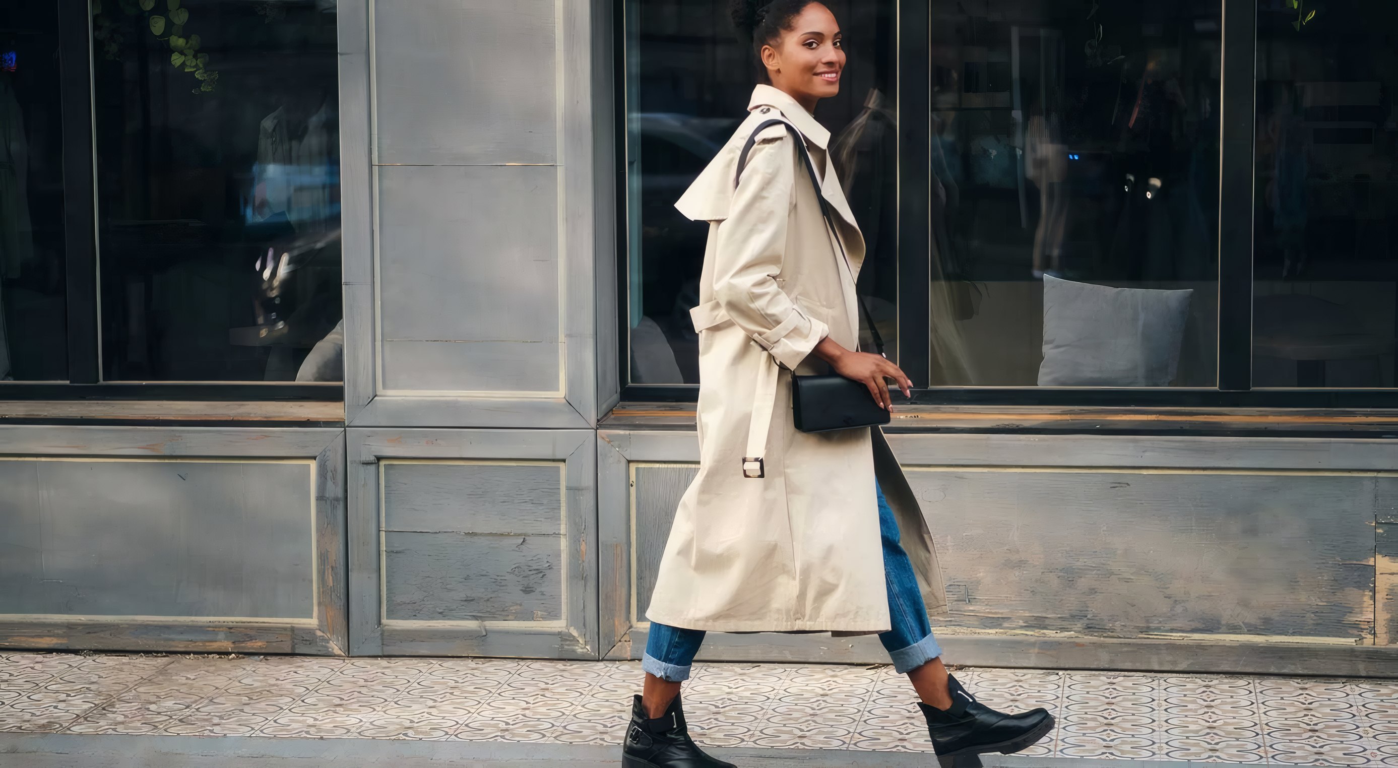 A woman in a trench coat and black boots walking past a shop front with a small bag