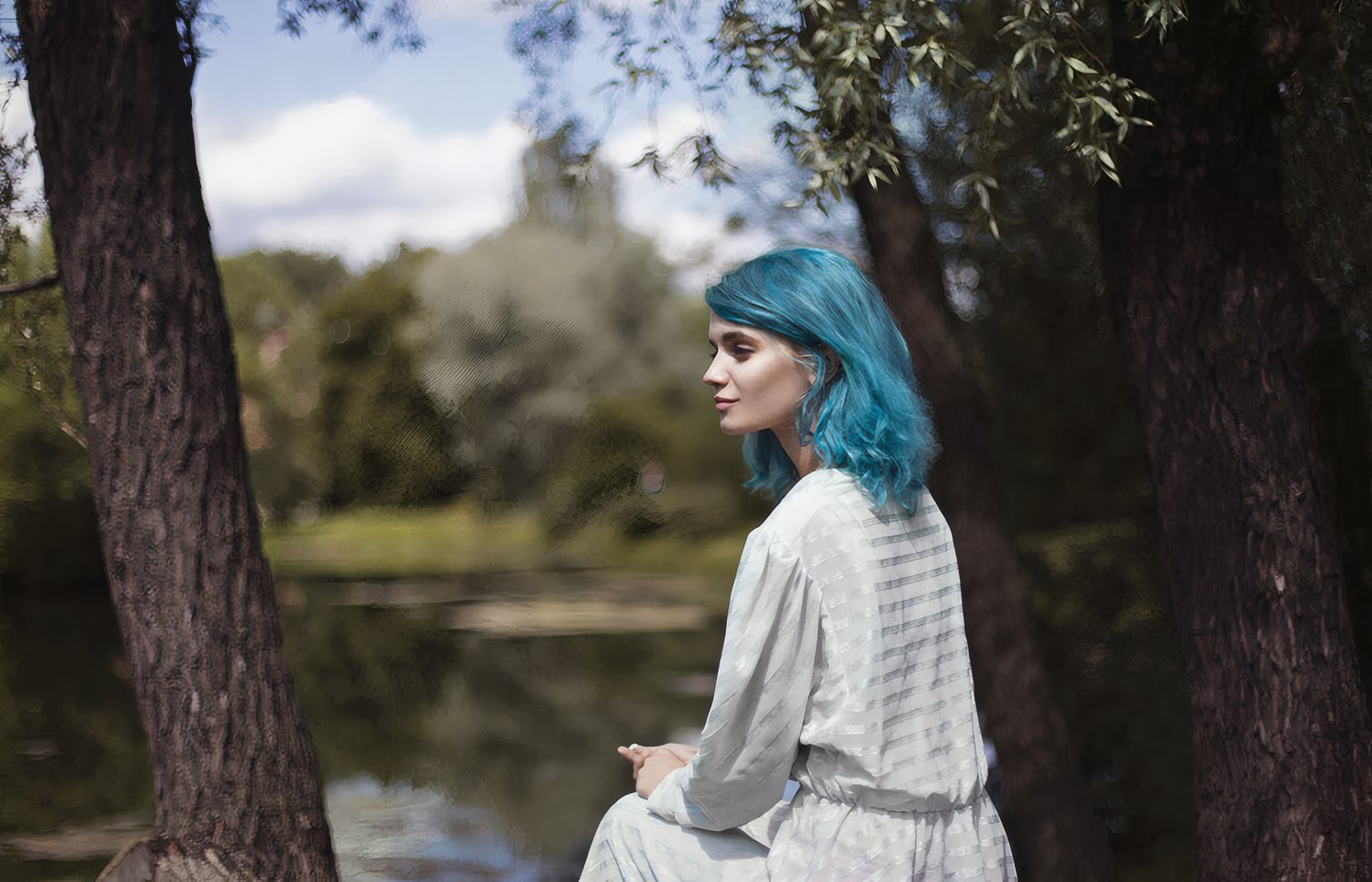 A woman with blue hair sitting by a river under trees