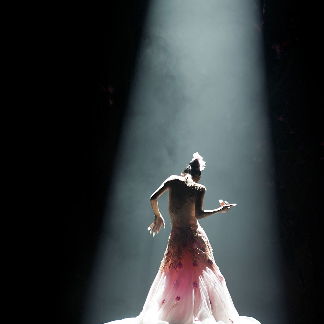 A dancer in a pale skirt under a single beam of stage light