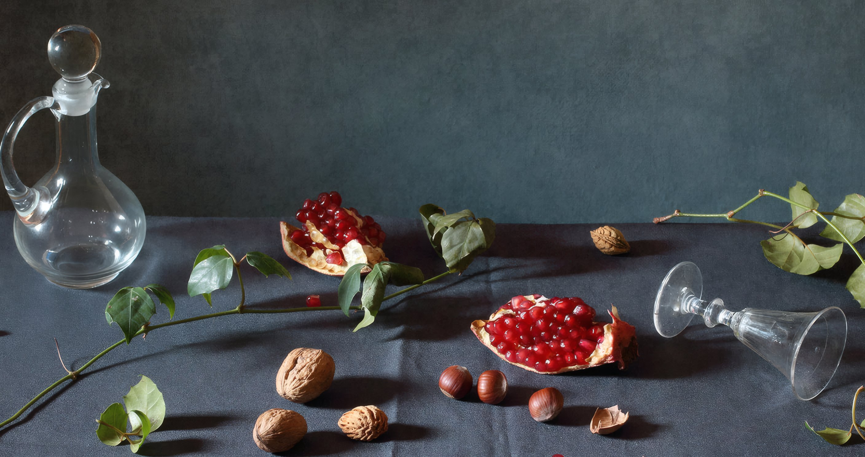 A glass carafe on its side among pomegranates and walnuts on a blue cloth