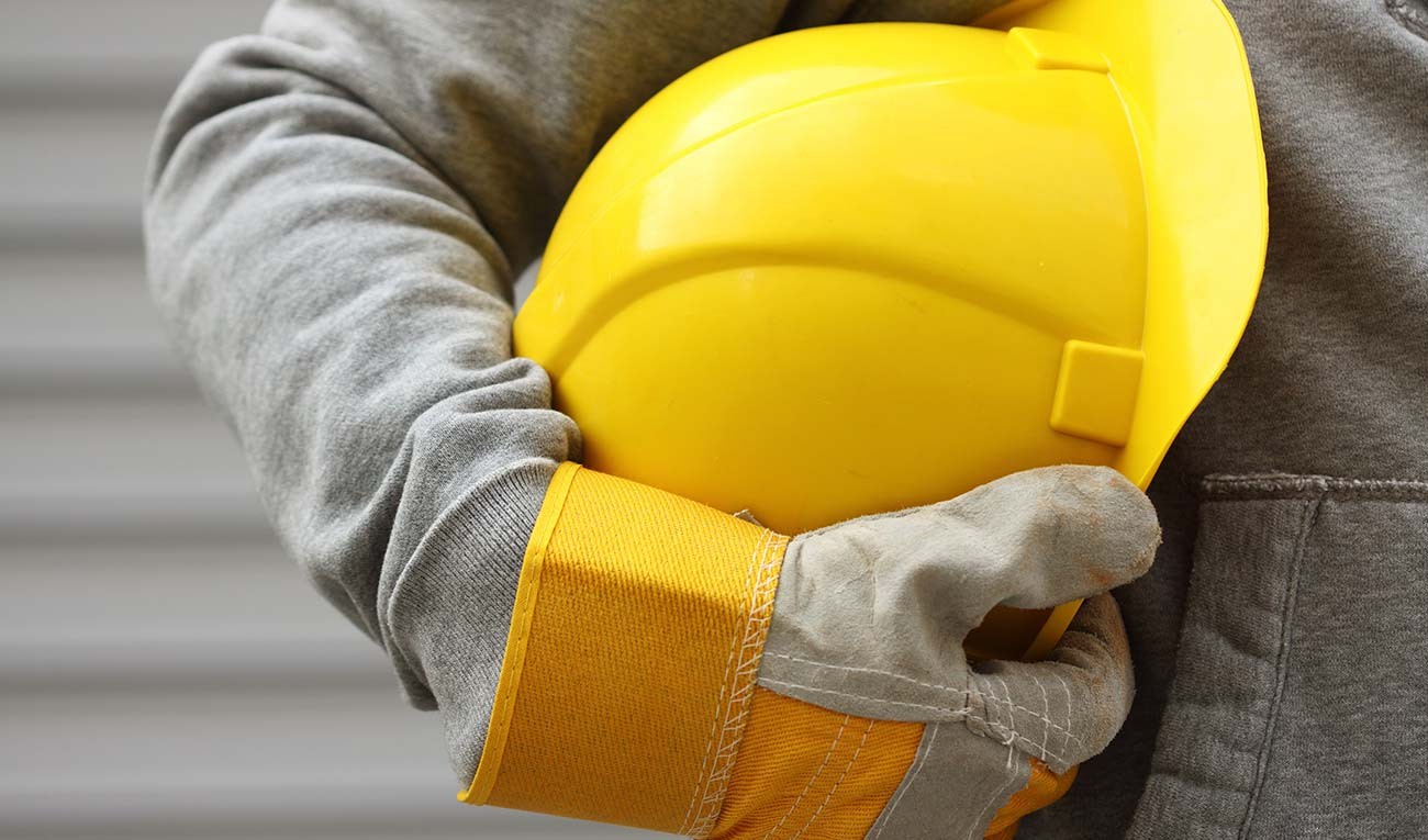 A hand in a work glove holding a yellow hard hat