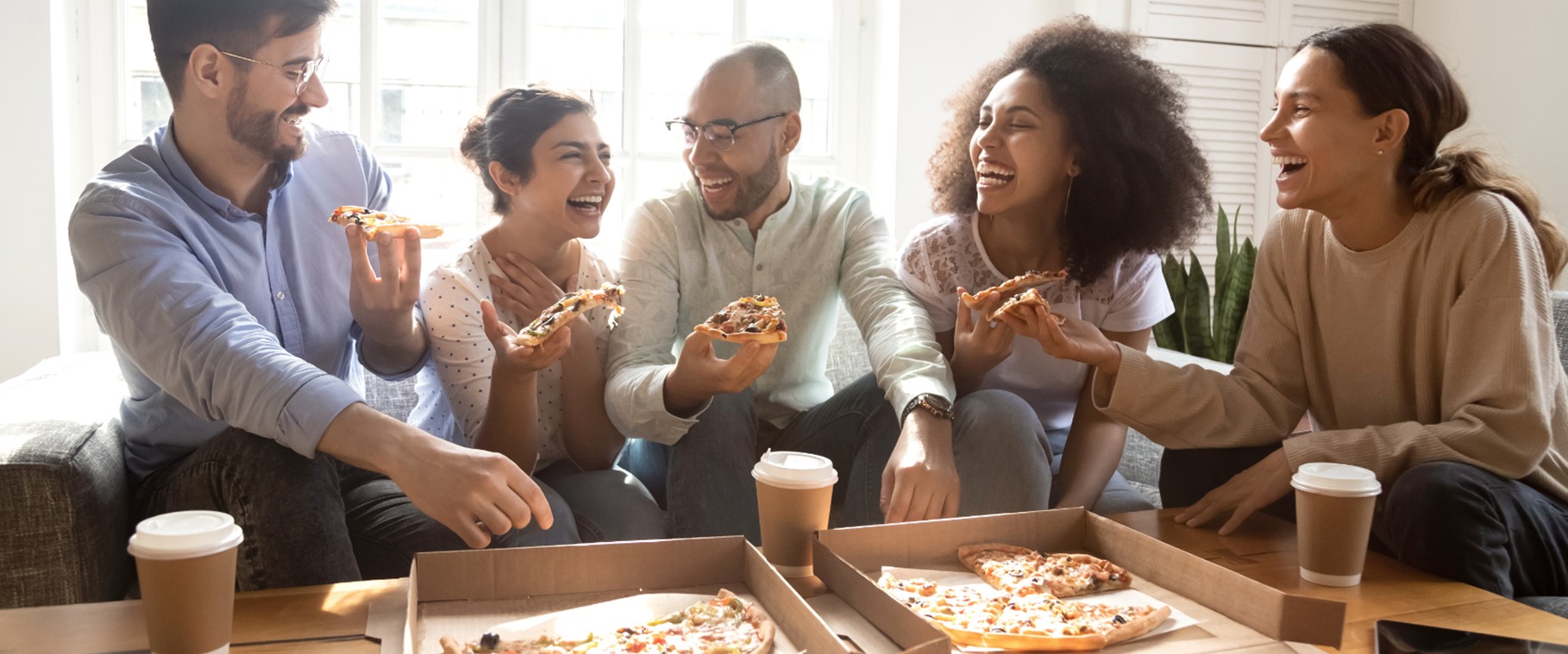 Five friends laughing round open pizza boxes on a sofa