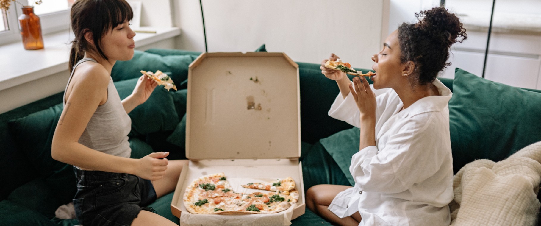 Two friends sharing slices from a box on a green sofa