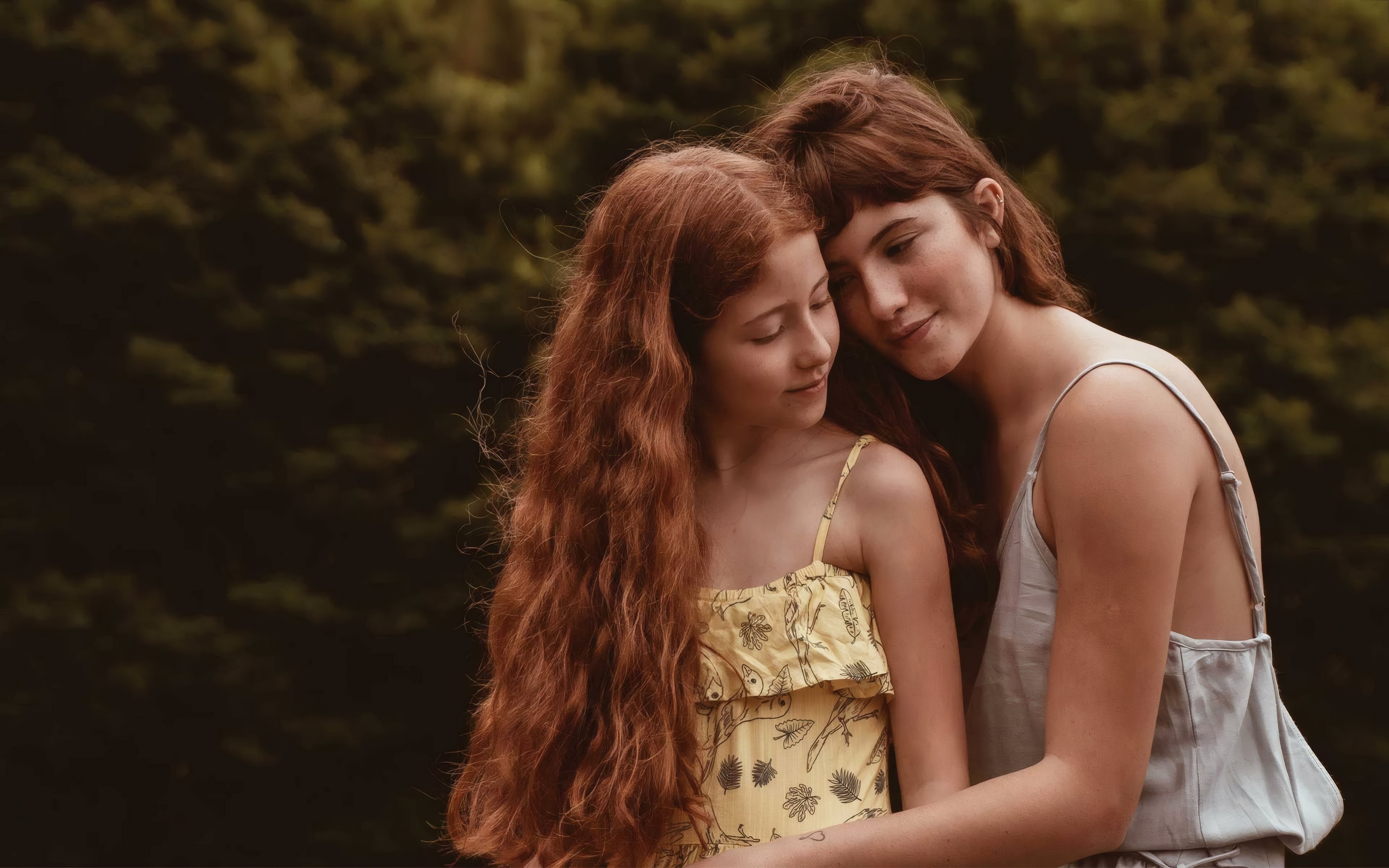 Two sisters with long red hair leaning together under the trees
