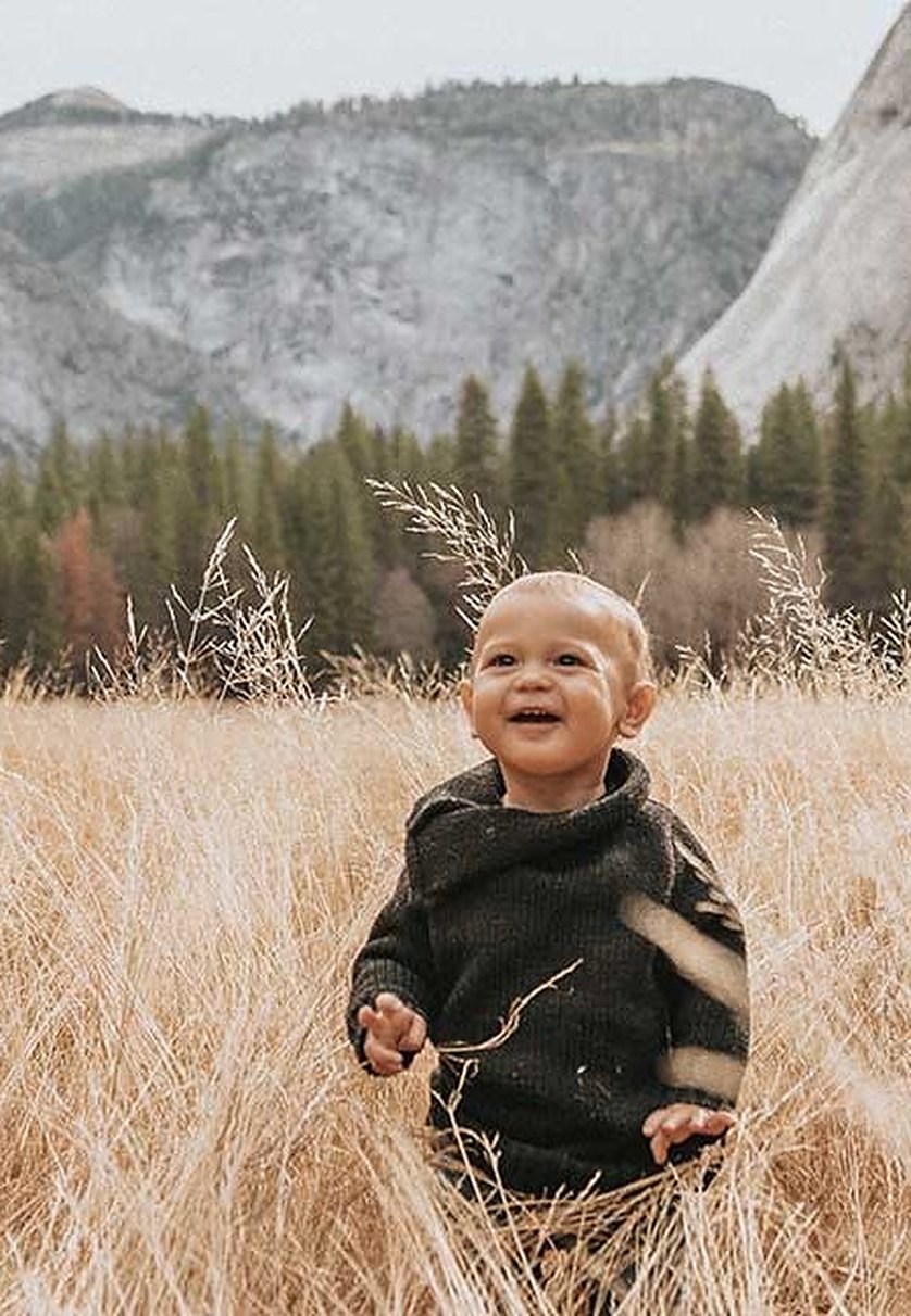 A toddler in a dark knit walking through tall golden grass