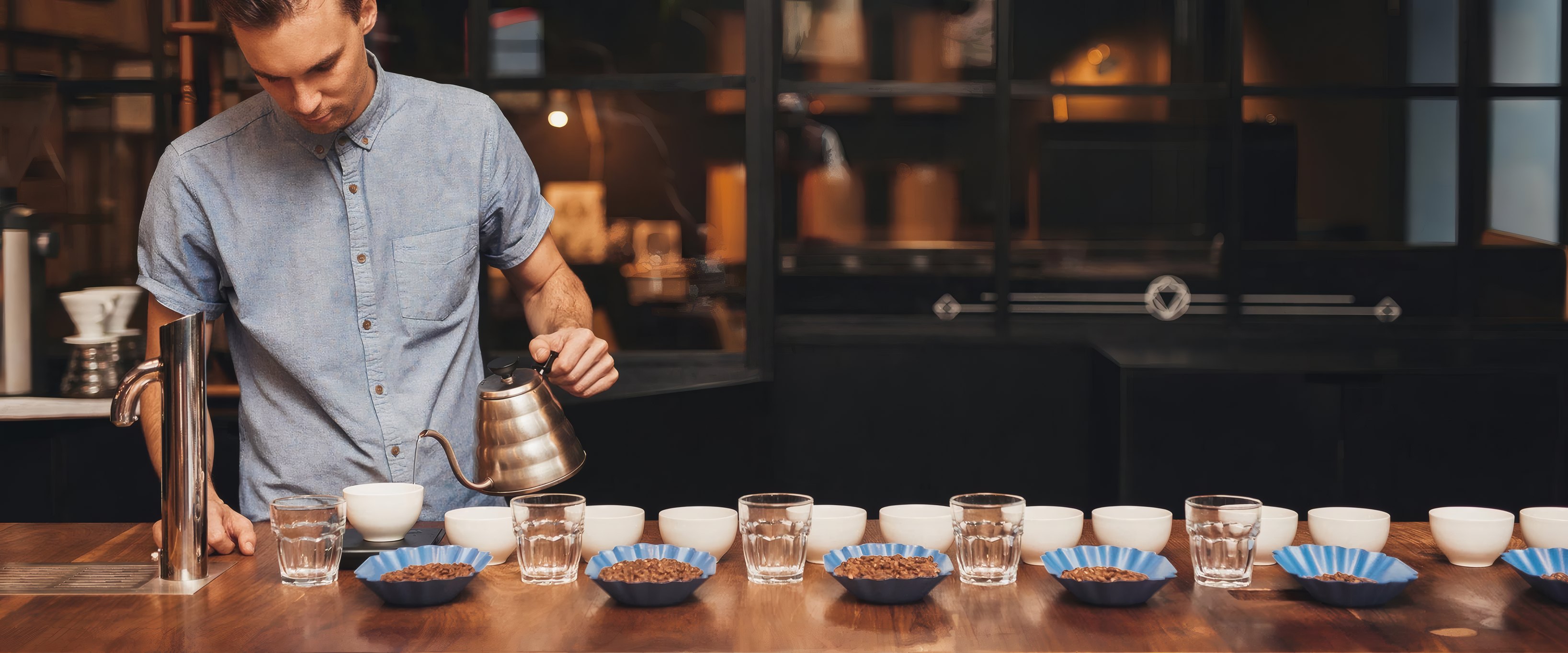 A barista pouring filter coffee over a row of cupping bowls