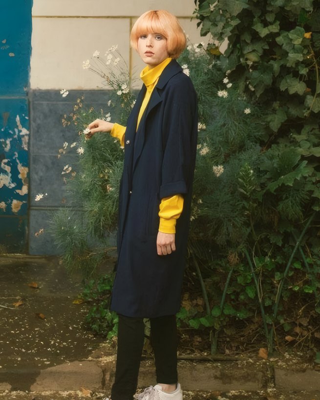 A long navy coat over a mustard jumper by a blue wall