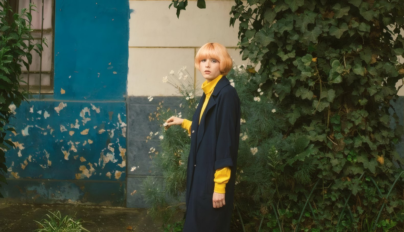 A woman in a long navy coat and a mustard jumper by an ivy-covered wall