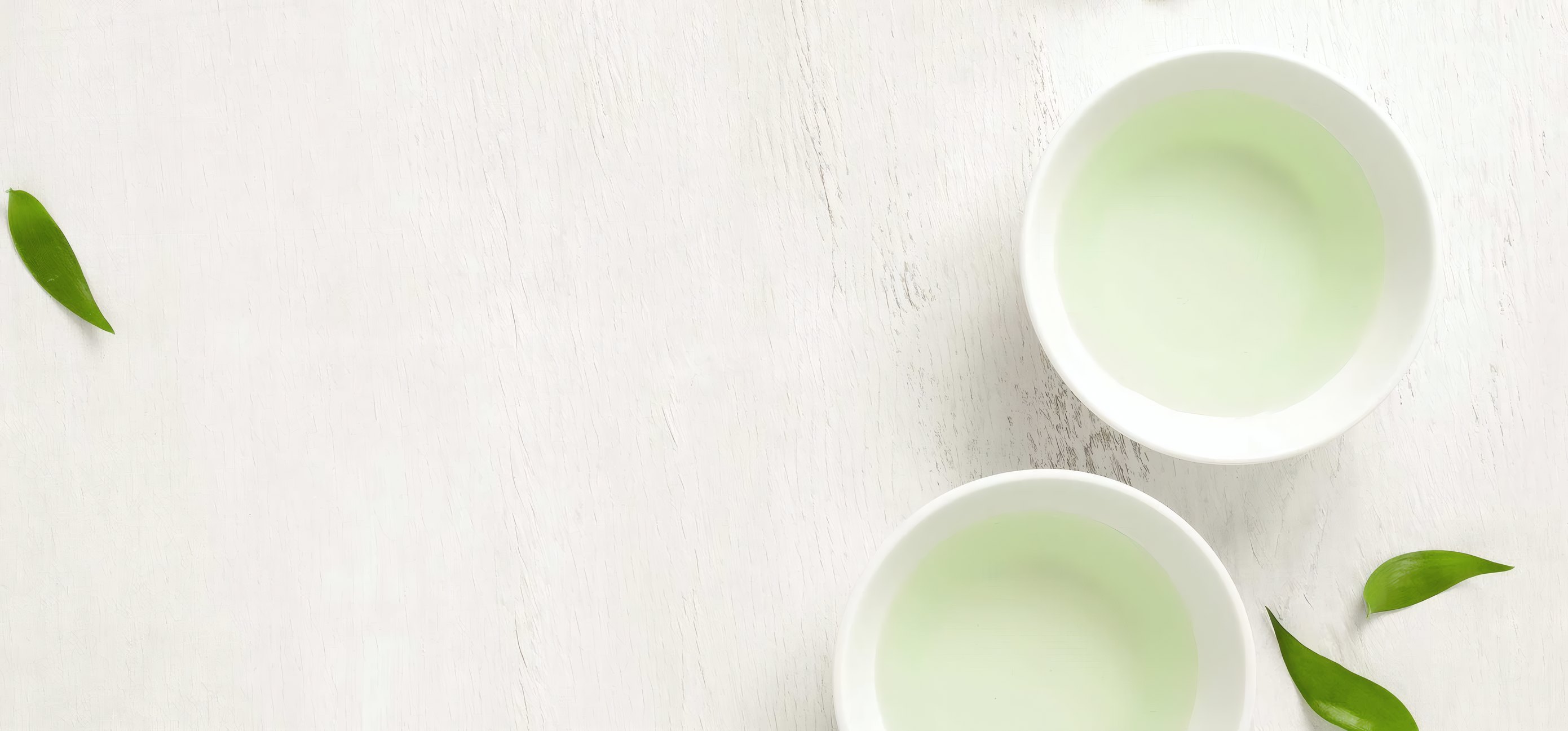 Two bowls of pale green tea and fresh leaves on a white table